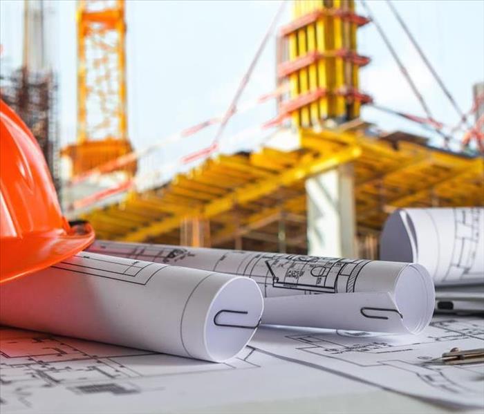A hard hat and building plans are on a table in front of a construction site.